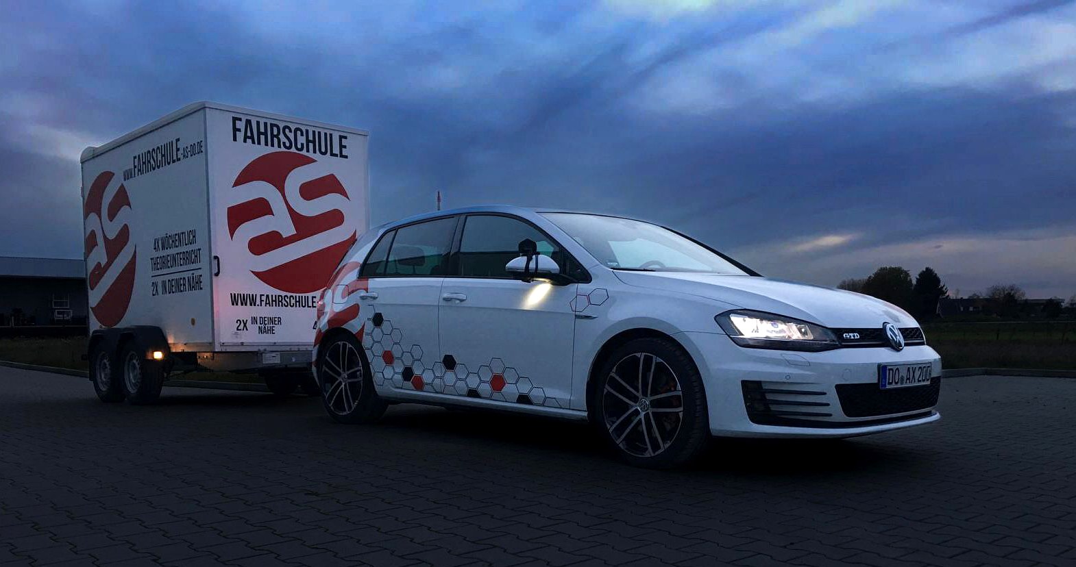 Ein foliertes Fahrschulfahrzeug der Fahrschule AS mit dem eigenen Werbeanhänger, abends bei Dämmerung auf einem Platz