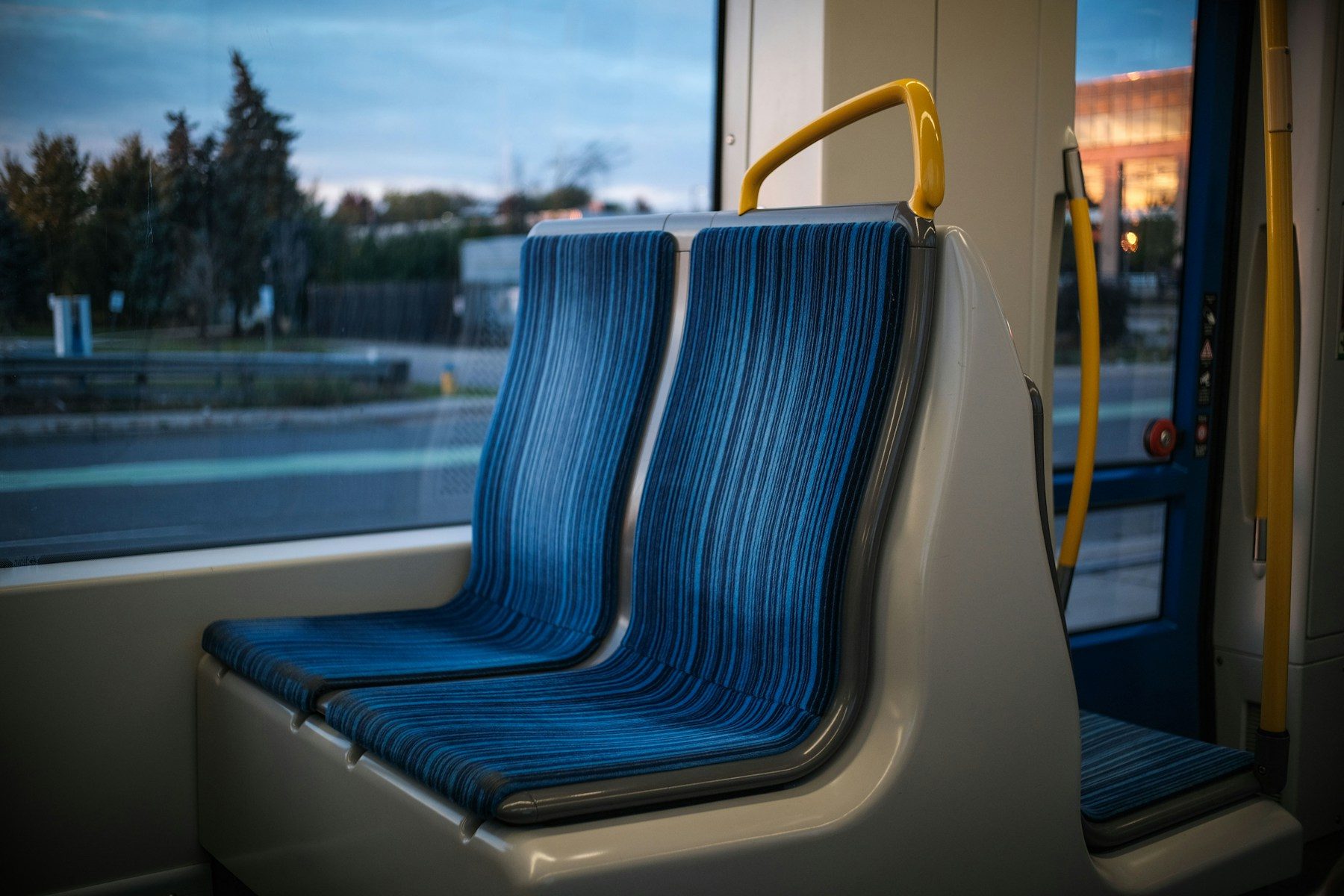 Leere blaue Doppelsitzbank am Fenster eines Busses, gelber Haltegriff, draußen Dämmerungslicht