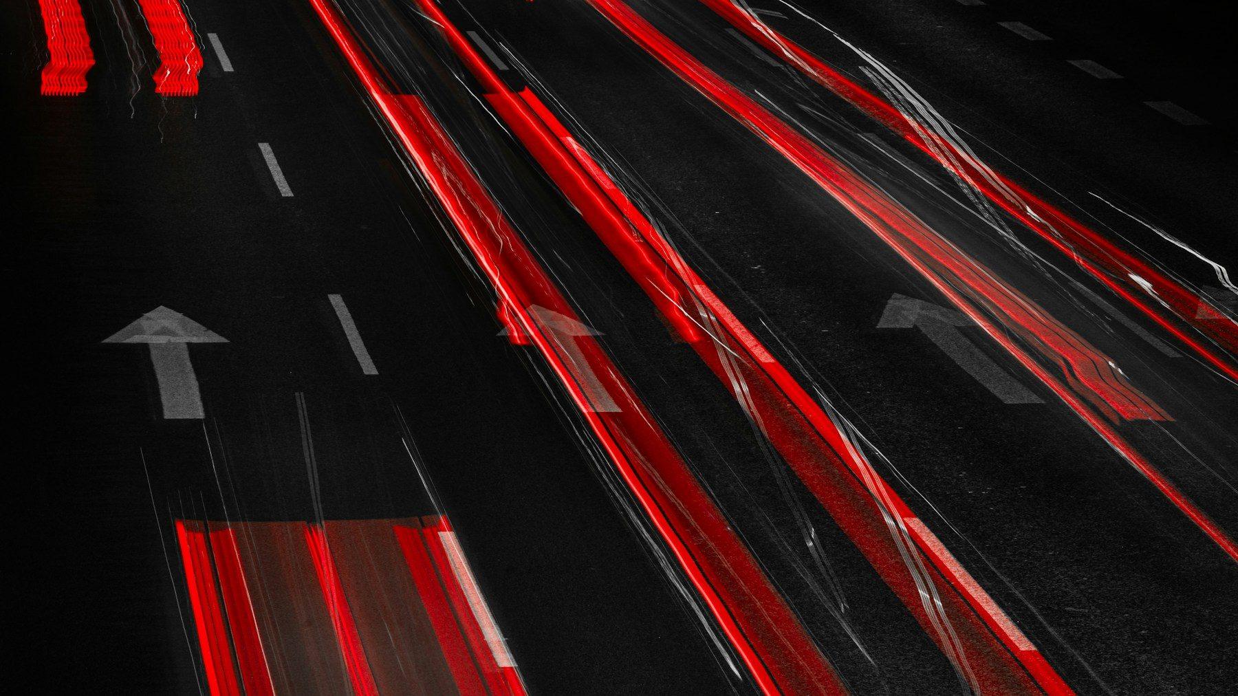 Nächtliche Fahrbahn von oben, rote Rücklicht-Streifen einer Langzeitbelichtung über dunklem Asphalt